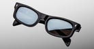 The Jacques Marie Mage Initials sunglasses by Jacques Marie Mage feature a black rectangular design with thick frames and blue-tinted lenses, set against a light gray backdrop—a sleek limited-edition eyewear choice.