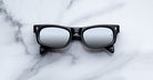 A pair of Jacques Marie Mage Initials black micro-frame sunglasses with mirrored lenses is placed on a white marble surface.
