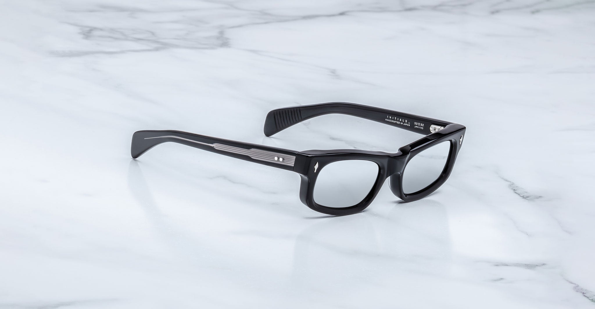 Jacques Marie Mage Initials black rectangular eyeglasses with thick frames, inspired by the brand's limited-edition designs, rest on a white marble surface.