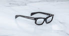 Jacques Marie Mage Initials black rectangular eyeglasses with thick frames, inspired by the brand's limited-edition designs, rest on a white marble surface.