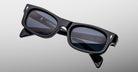The Jacques Marie Mage Initials by Jacques Marie Mage are black micro-frame smart glasses with rectangular lenses, displaying digital text and icons on the right lens against a plain light background.