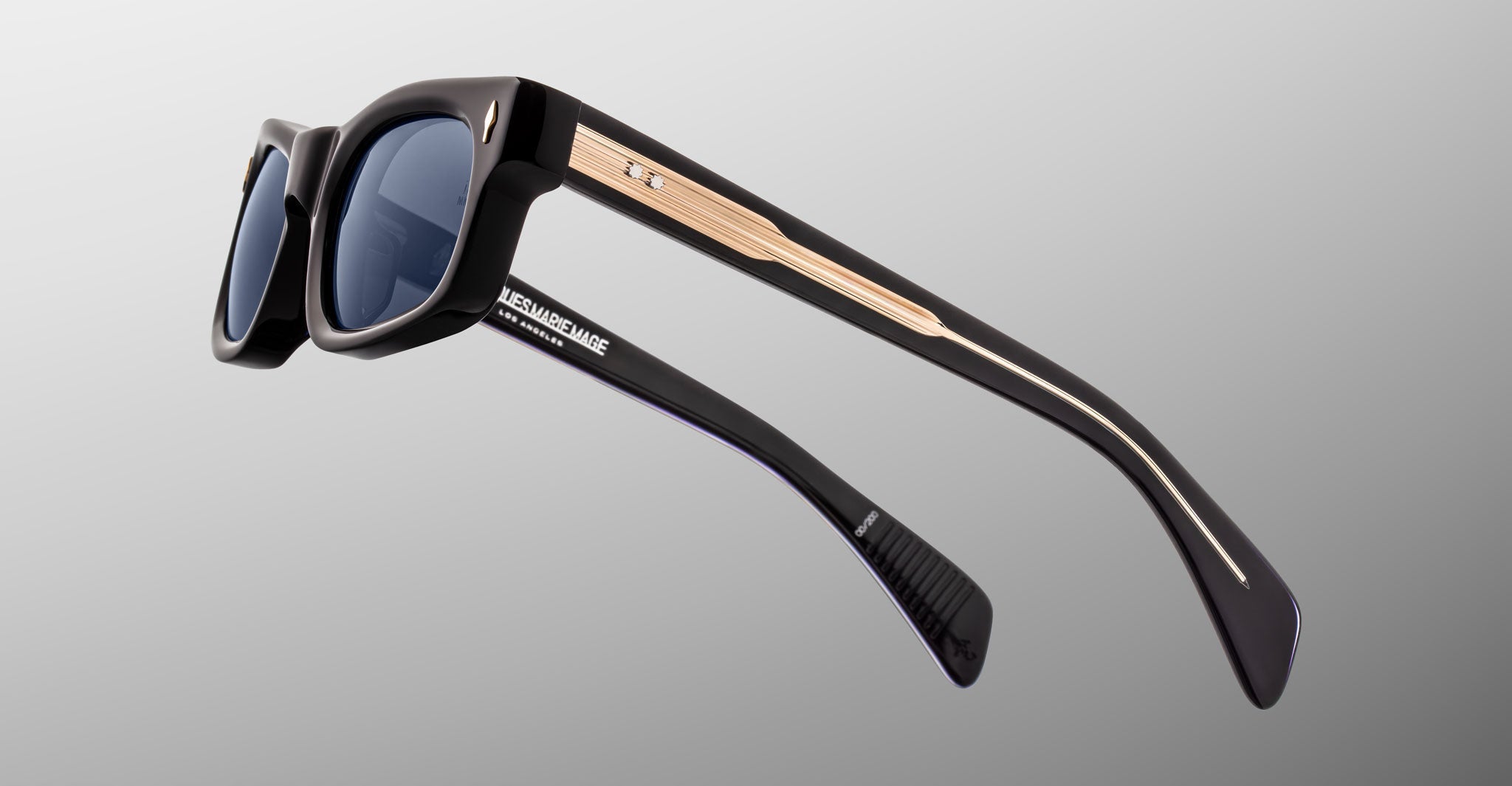 Jacques Marie Mage Initials sunglasses in black with blue lenses and gold accents on the arms, featuring visible Jacques Marie Mage branding inside the left arm. This limited-edition style is set against a light gray background.