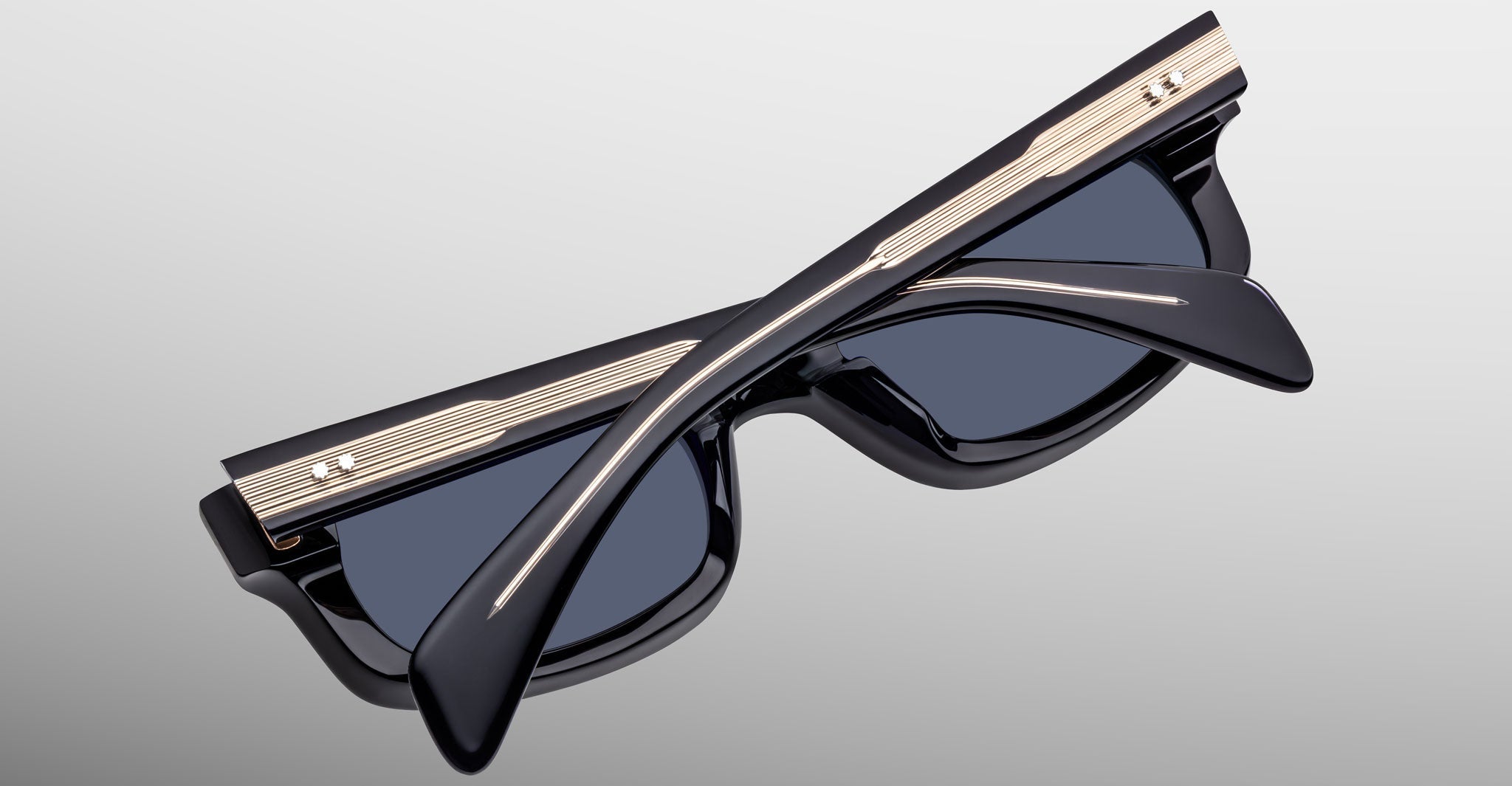 A pair of black Jacques Marie Mage Initials sunglasses with gold accents, limited-edition frames by Jacques Marie Mage, is folded closed and set on a light gray surface.