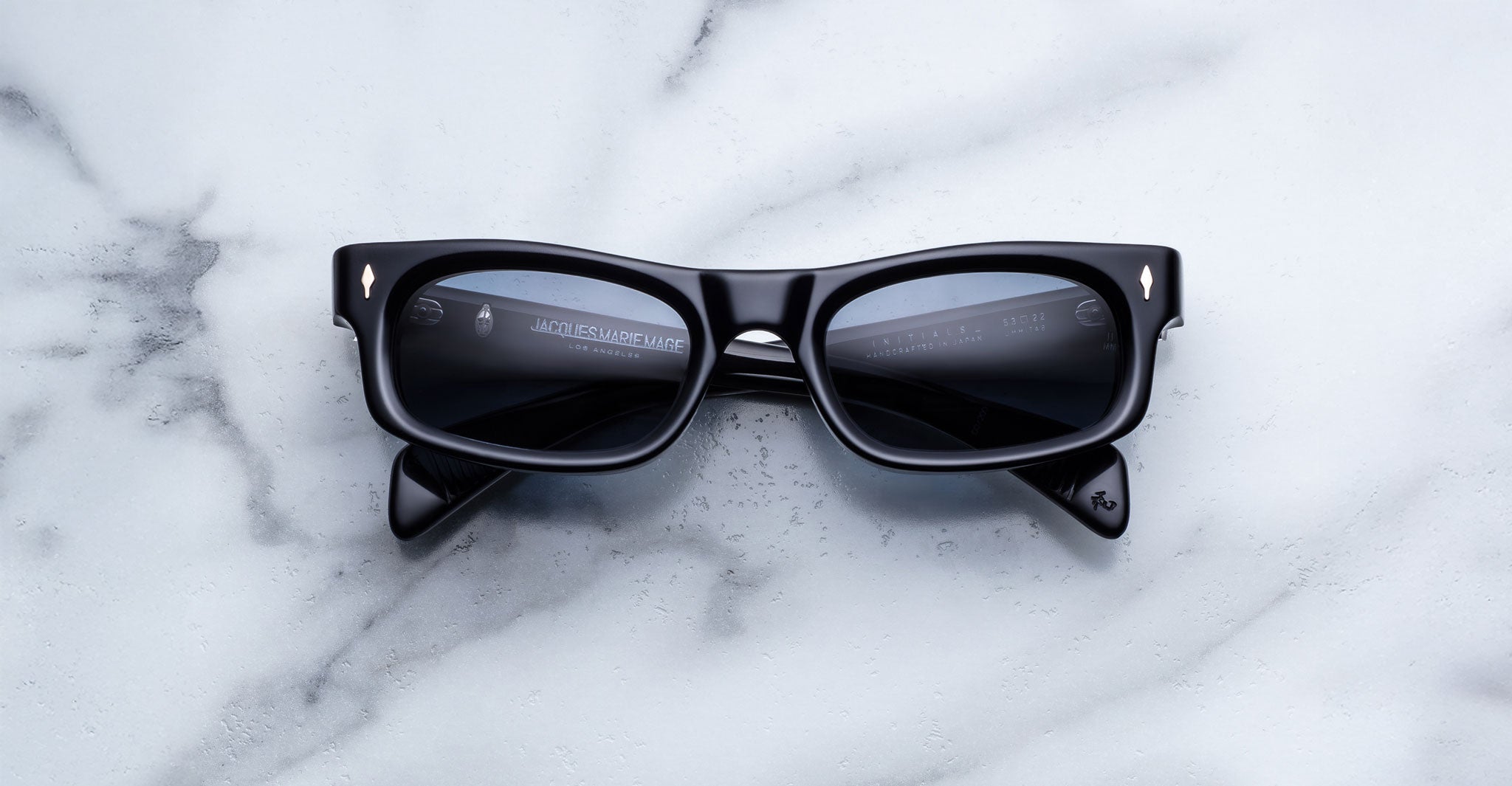 Jacques Marie Mage Initials black rectangular limited-edition sunglasses with thick frames are displayed on a white marble surface.
