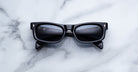 Jacques Marie Mage Initials black rectangular limited-edition sunglasses with thick frames are displayed on a white marble surface.