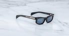 Jacques Marie Mage Initials: Black rectangular micro-frame sunglasses with dark lenses and metallic arm accents, shown on a white marble surface. Limited-edition frames inspired by the signature Jacques Marie Mage style.