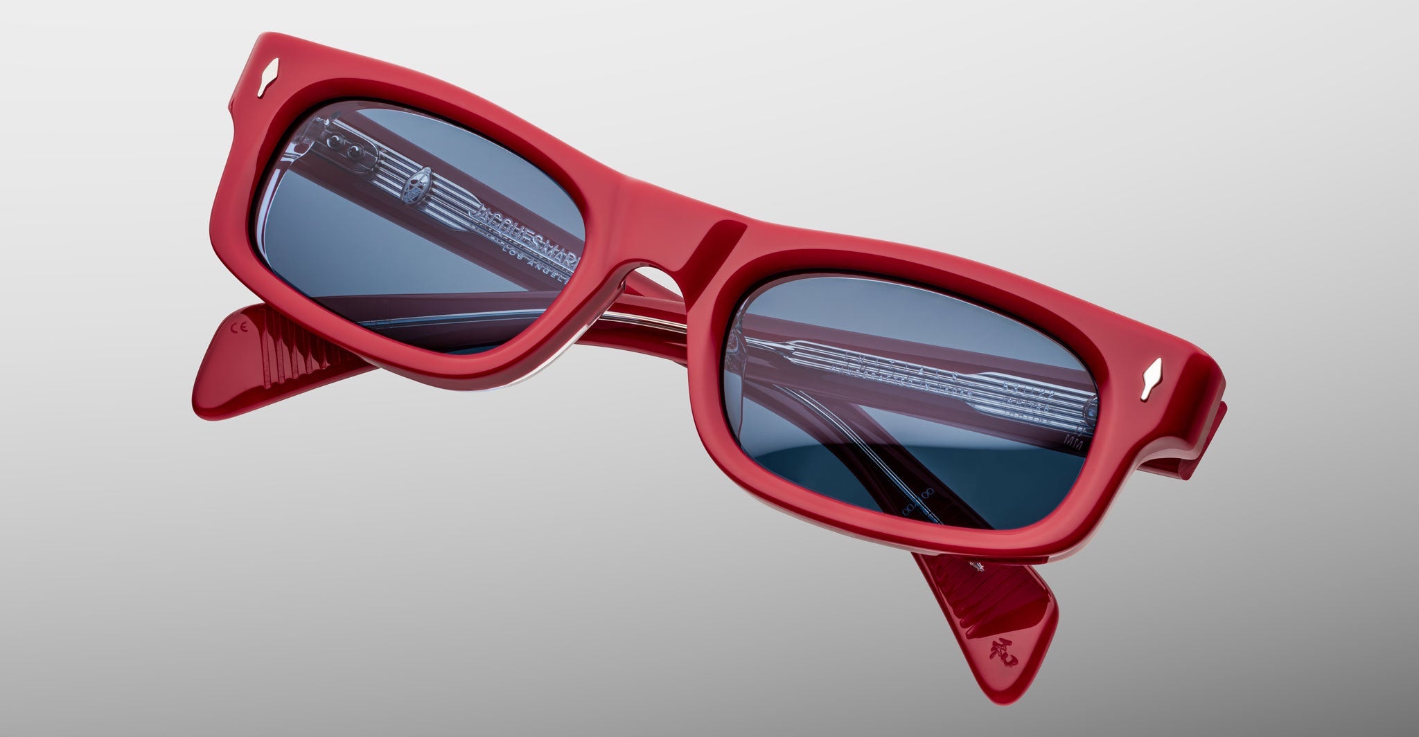 The Jacques Marie Mage Initials sunglasses feature red micro-frames and dark blue lenses, showcased here against a light gray background. This limited-edition design reflects the signature style of Jacques Marie Mage.