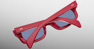 A pair of limited-edition Jacques Marie Mage Initials sunglasses with red micro-frames and blue-tinted lenses, folded and displayed on a light gray surface.