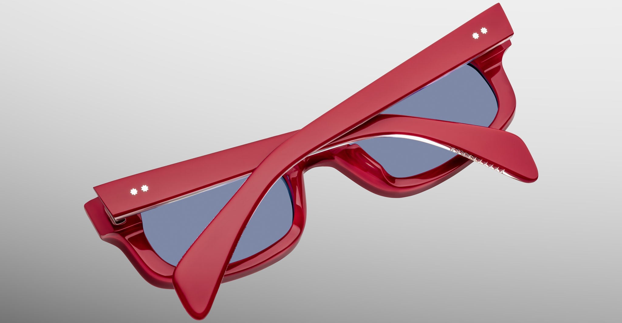 A pair of limited-edition Jacques Marie Mage Initials sunglasses with red micro-frames and blue-tinted lenses, folded and displayed on a light gray surface.