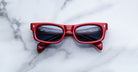 Jacques Marie Mage Initials in Pirate red-framed sunglasses with dark lenses, folded, viewed from above.