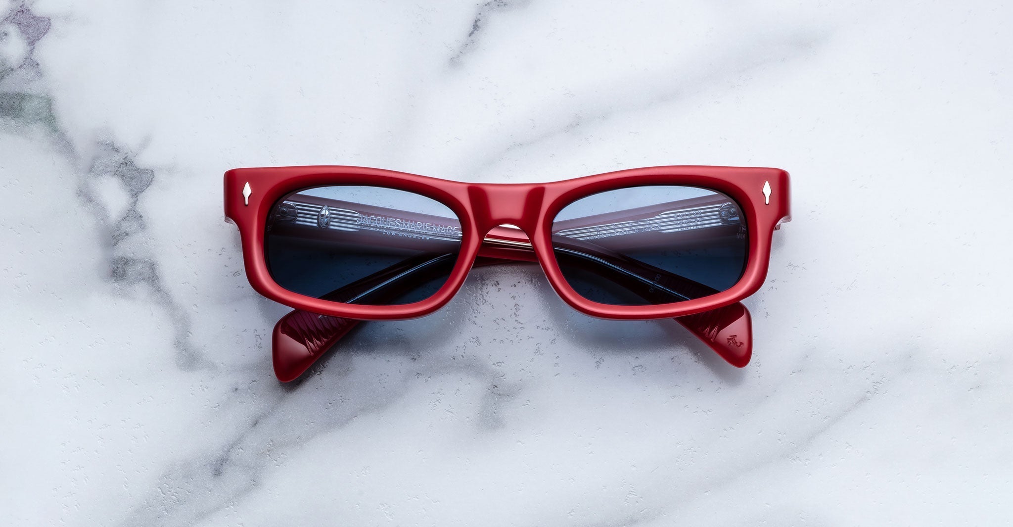 Jacques Marie Mage Initials micro-frame sunglasses with red frames and dark lenses are folded on a white marble surface.