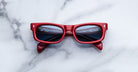 Jacques Marie Mage Initials micro-frame sunglasses with red frames and dark lenses are folded on a white marble surface.