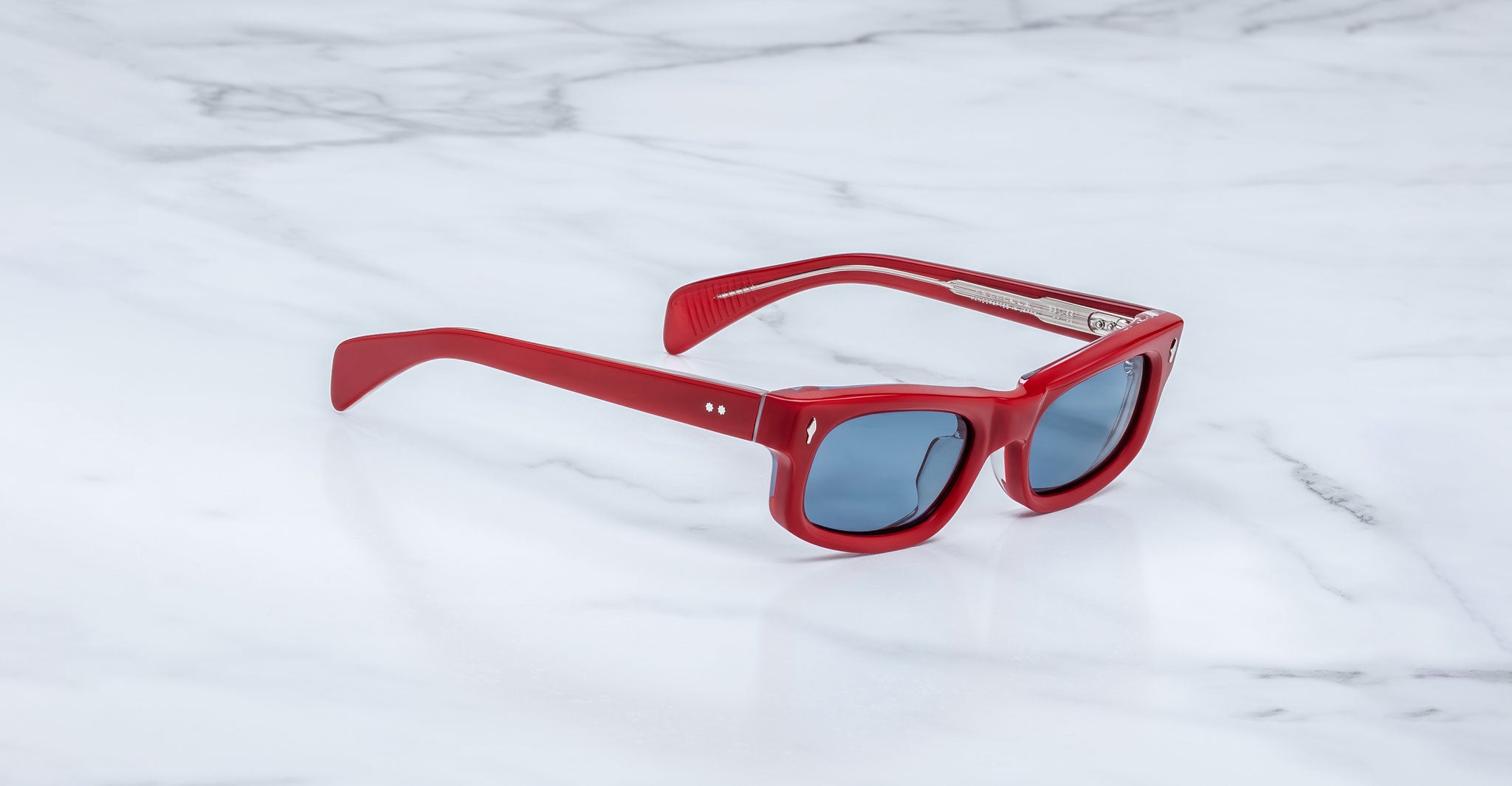 Jacques Marie Mage Initials sunglasses with red frames and dark lenses rest on a white marble surface, showcasing the brand's exclusive limited-edition style.