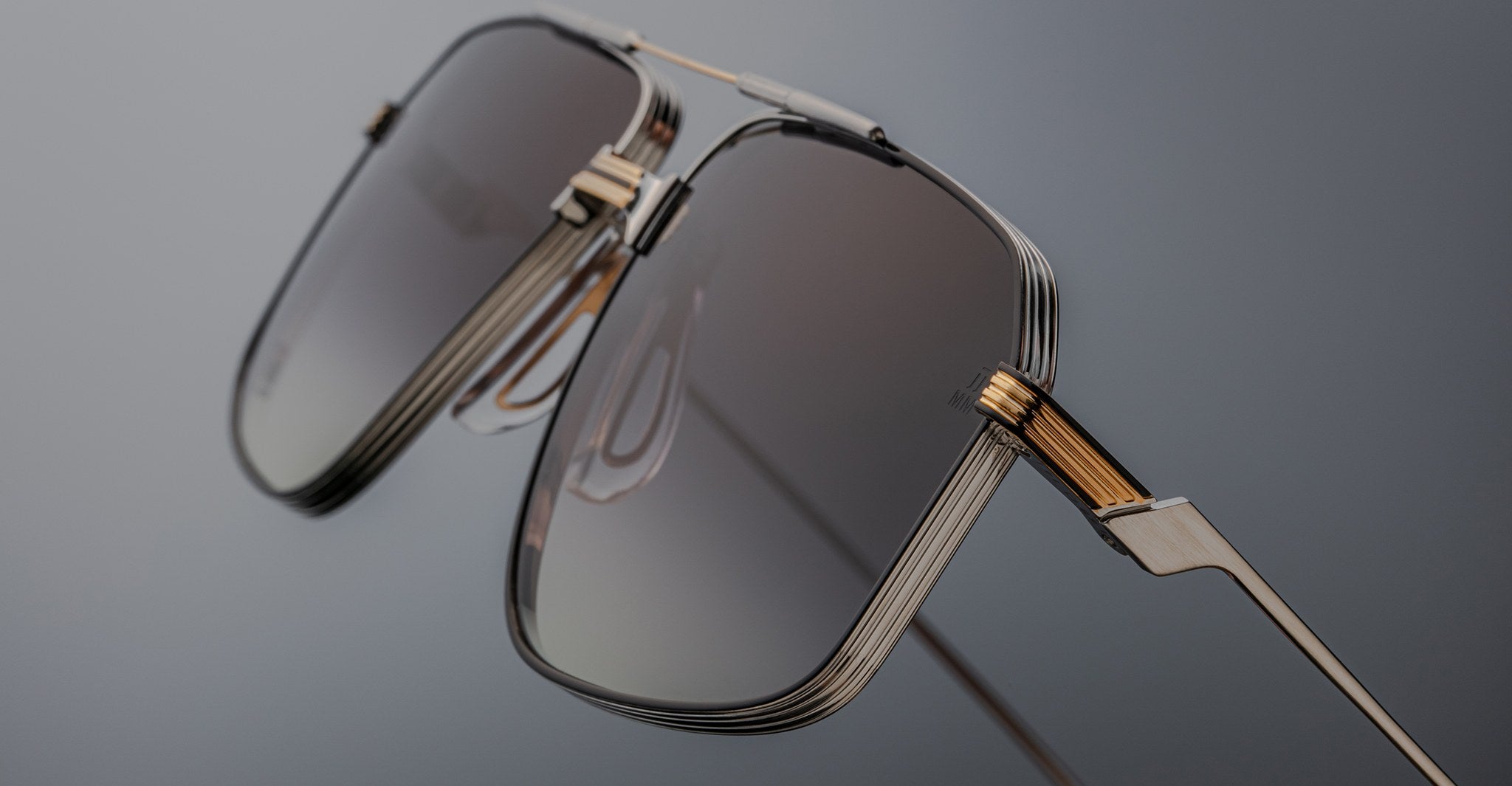 Close-up of Jacques Marie Mage Jagger sunglasses by Jacques Marie Mage, showcasing oversized aviator style with gold and silver beta titanium frames and rectangular gradient tinted lenses against a gray background.