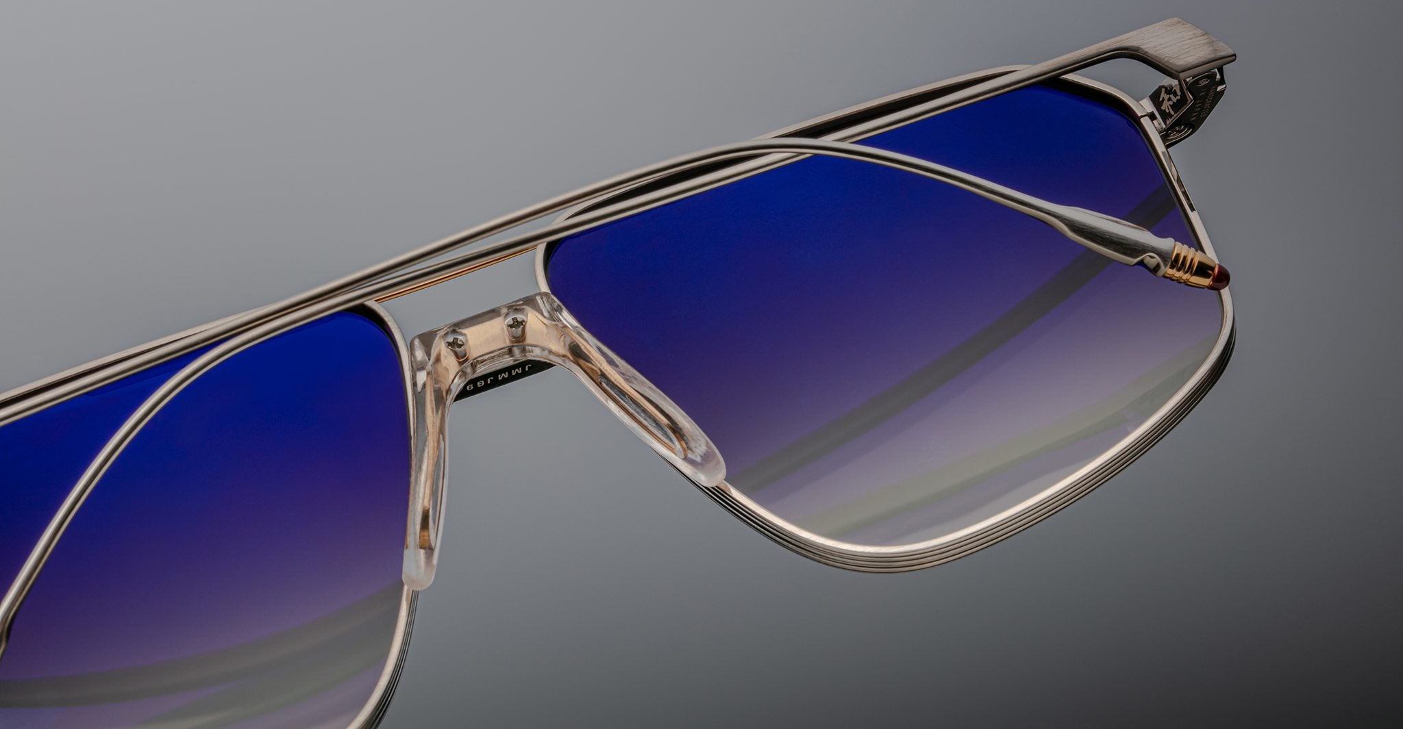 Close-up of Jacques Marie Mage Jagger oversized aviator sunglasses by Jacques Marie Mage, featuring beta titanium metallic frames and blue gradient lenses against a plain gray background.