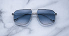 A pair of Jacques Marie Mage Jagger oversized aviator sunglasses with blue lenses and a sleek silver beta titanium frame by Jacques Marie Mage, displayed on a white marble surface.