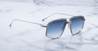 Jacques Marie Mage Jagger oversized aviator sunglasses with a silver frame and blue gradient lenses rest on white marble, channeling the iconic style of Jacques Marie Mage.