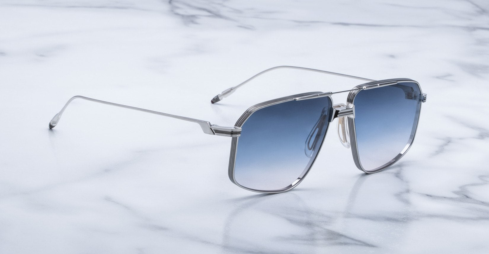 Jacques Marie Mage Jagger oversized aviator sunglasses with a silver frame and blue gradient lenses rest on white marble, channeling the iconic style of Jacques Marie Mage.