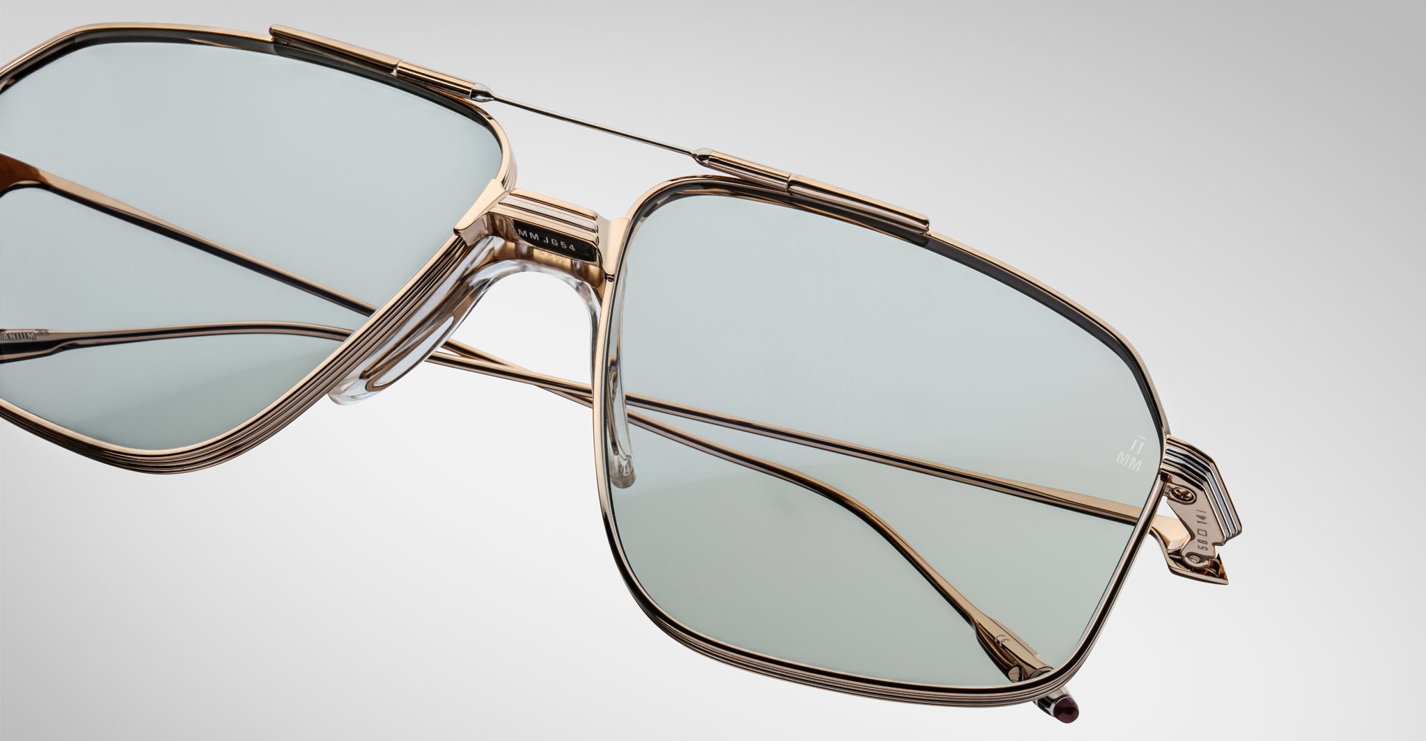 Close-up of the Jacques Marie Mage Jagger oversized aviator sunglasses by Jacques Marie Mage, with gold frames and light blue-tinted lenses set against a plain, gradient background.