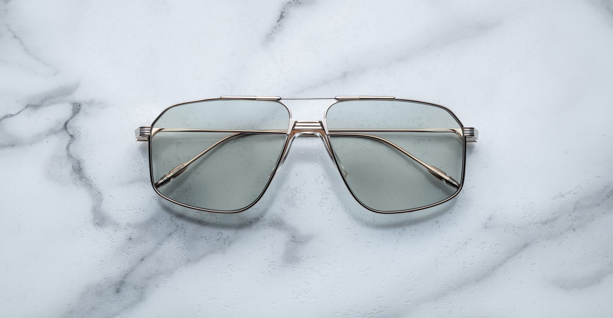 Jacques Marie Mage Jagger oversized aviator sunglasses by Jacques Marie Mage, featuring gold beta titanium frames and tinted lenses, are shown on a white marble surface.