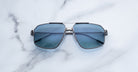 A pair of Jacques Marie Mage Jagger oversized aviator sunglasses with blue-tinted lenses and beta titanium frames by Jacques Marie Mage is displayed on a white marble surface.