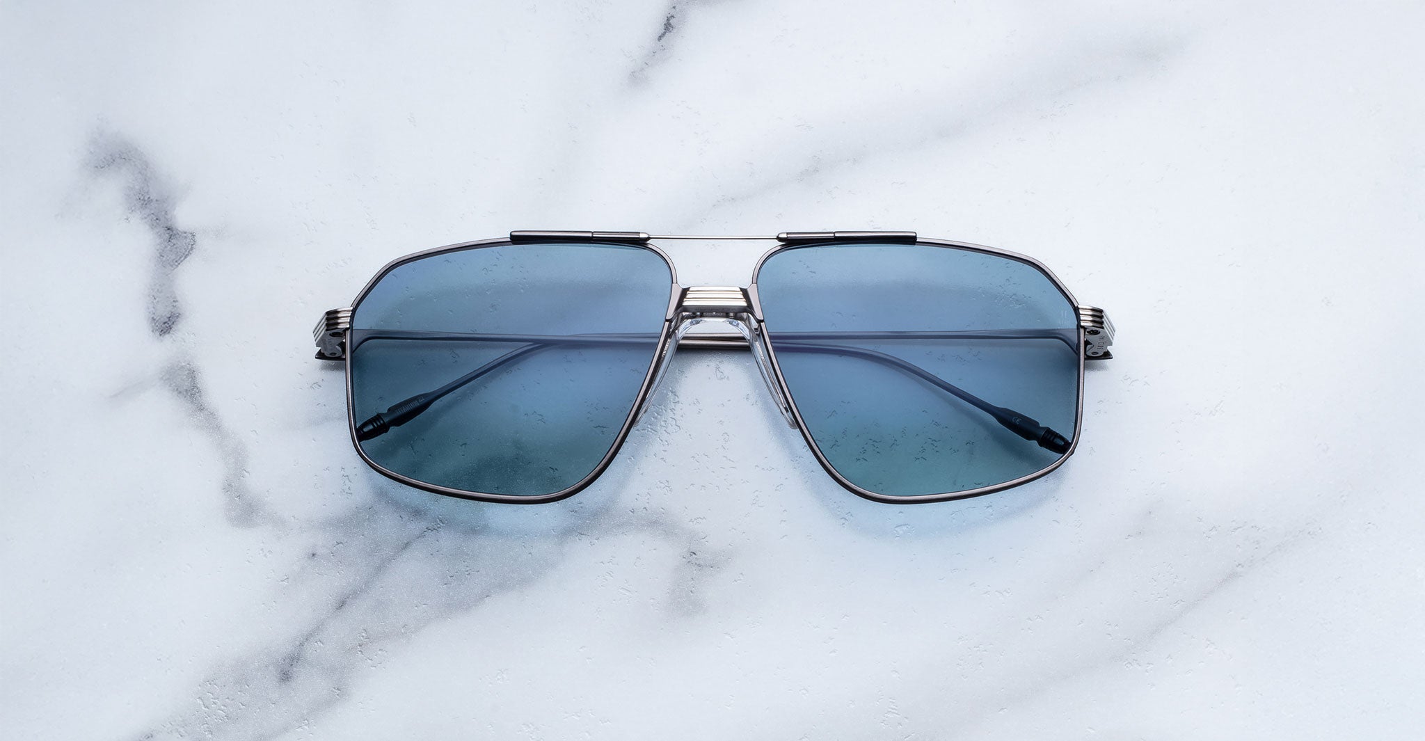 A pair of Jacques Marie Mage Jagger oversized aviator sunglasses with blue-tinted lenses and beta titanium frames by Jacques Marie Mage is displayed on a white marble surface.