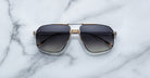 The Jacques Marie Mage Jagger sunglasses by Jacques Marie Mage, black oversized aviators with gold and beta titanium accents, are displayed on a white marble surface.