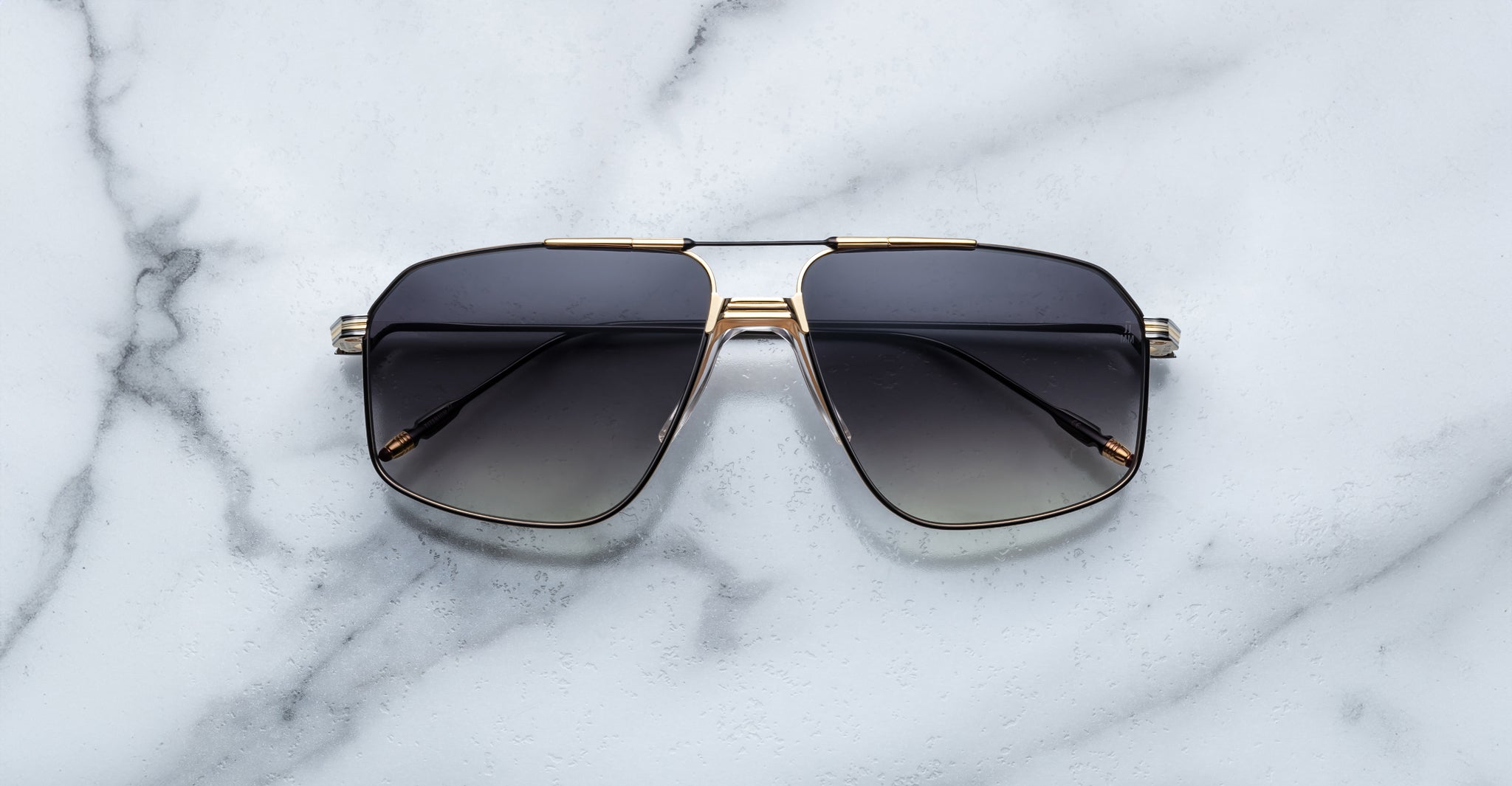 The Jacques Marie Mage Jagger sunglasses by Jacques Marie Mage, black oversized aviators with gold and beta titanium accents, are displayed on a white marble surface.