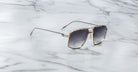 The Jacques Marie Mage Jagger sunglasses by Jacques Marie Mage feature oversized aviator frames with gold rims and dark lenses, exuding sophistication as they are elegantly displayed on a white marble surface.