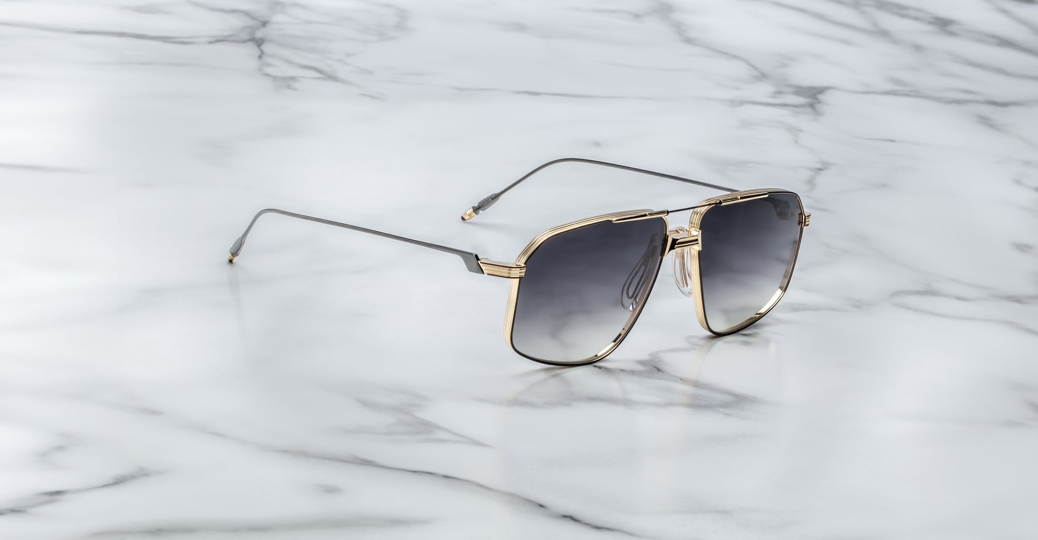 The Jacques Marie Mage Jagger sunglasses by Jacques Marie Mage feature oversized aviator frames with gold rims and dark lenses, exuding sophistication as they are elegantly displayed on a white marble surface.