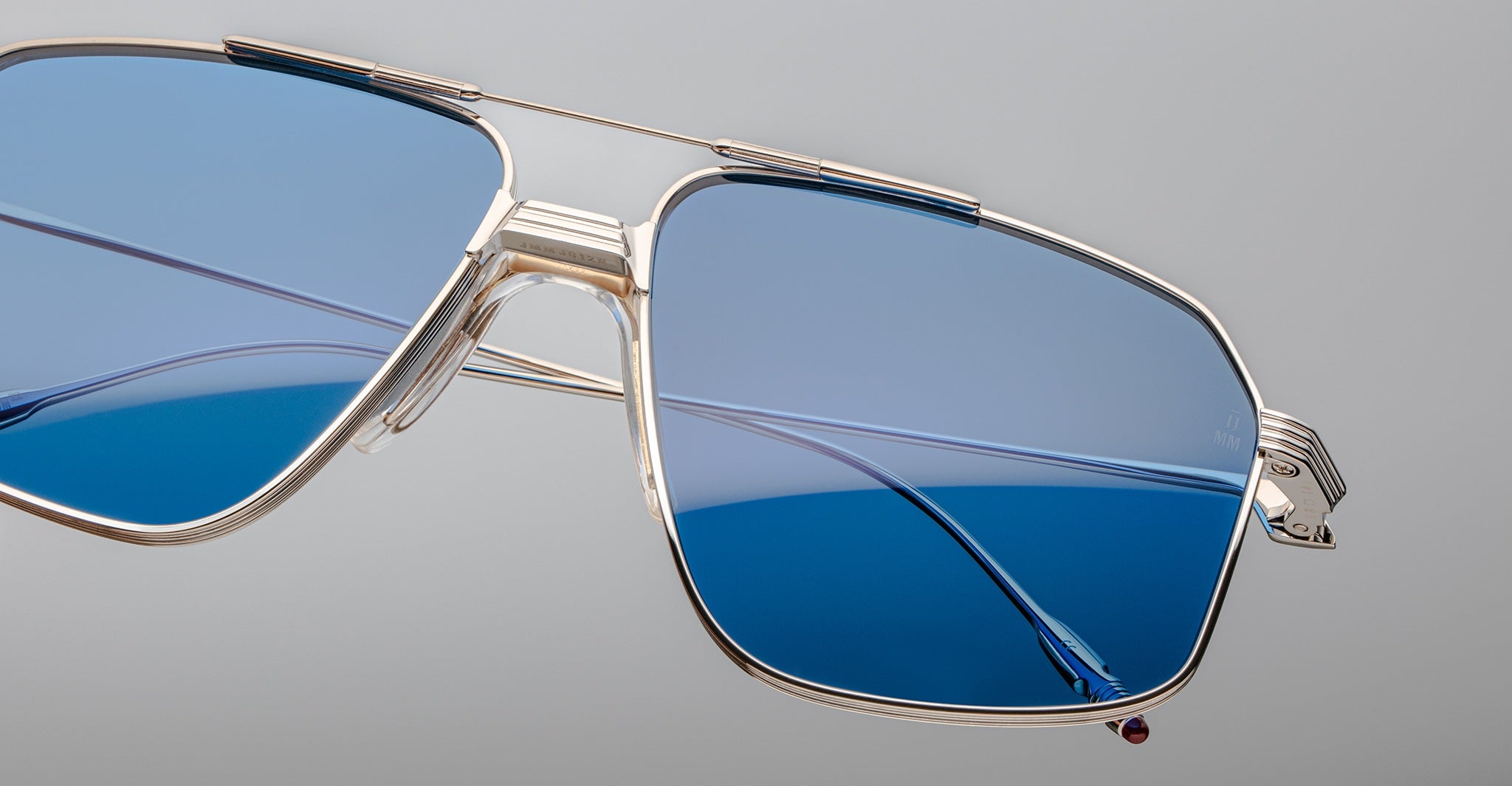 Close-up of Jacques Marie Mage Jagger oversized aviator sunglasses by Jacques Marie Mage, featuring gold metal frames, blue-tinted lenses, a double bridge, and lightweight beta titanium construction, shown against a plain grey background.