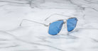 A pair of Jacques Marie Mage Jagger aviator sunglasses with silver frames and blue lenses rests on white marble, highlighting their sleek beta titanium design.