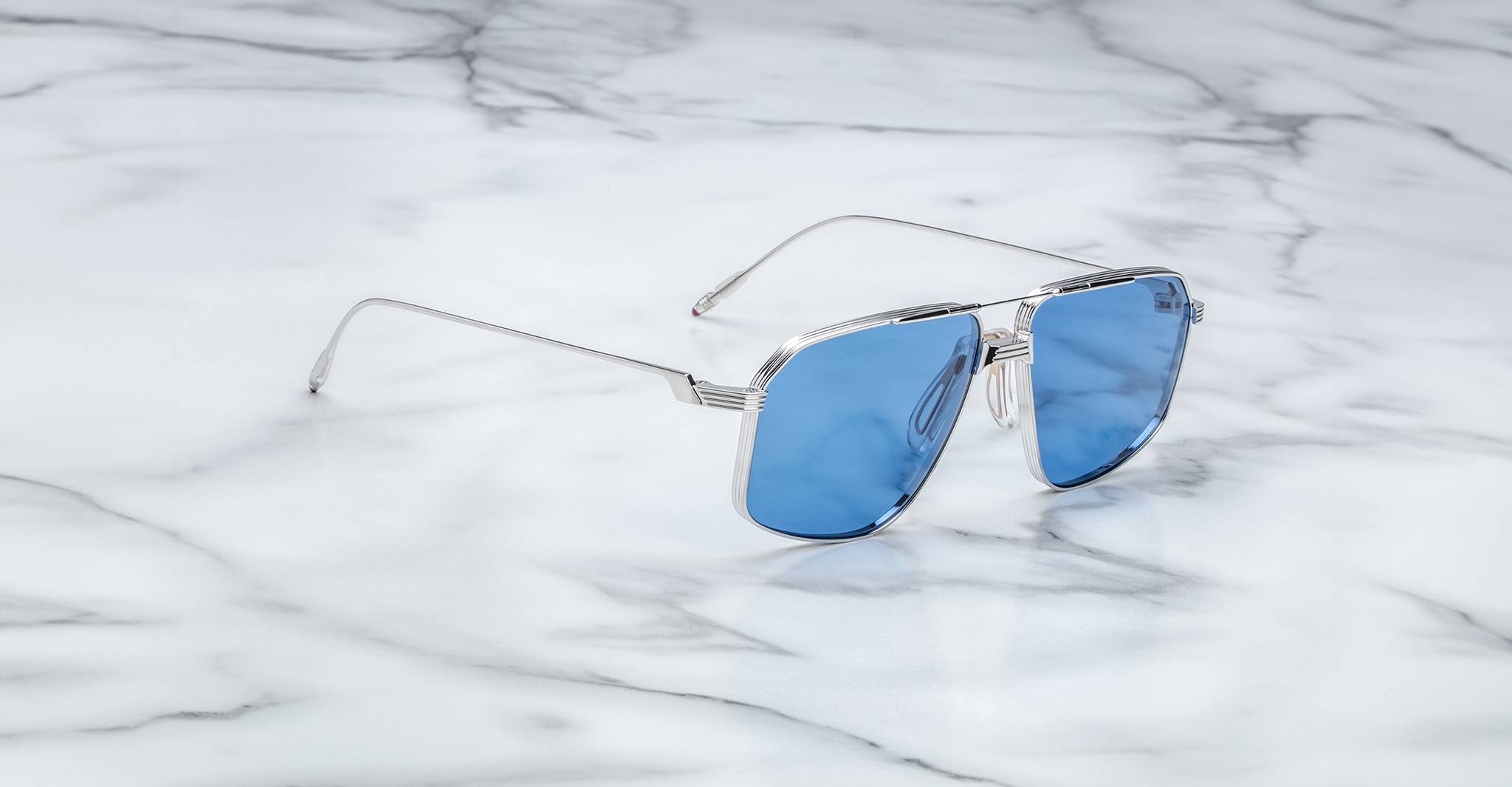 A pair of Jacques Marie Mage Jagger aviator sunglasses with silver frames and blue lenses rests on white marble, highlighting their sleek beta titanium design.