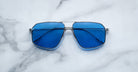 The Jacques Marie Mage Jagger sunglasses by Jacques Marie Mage feature oversized aviator frames in silver beta titanium with blue-tinted lenses, displayed on a white marble surface.
