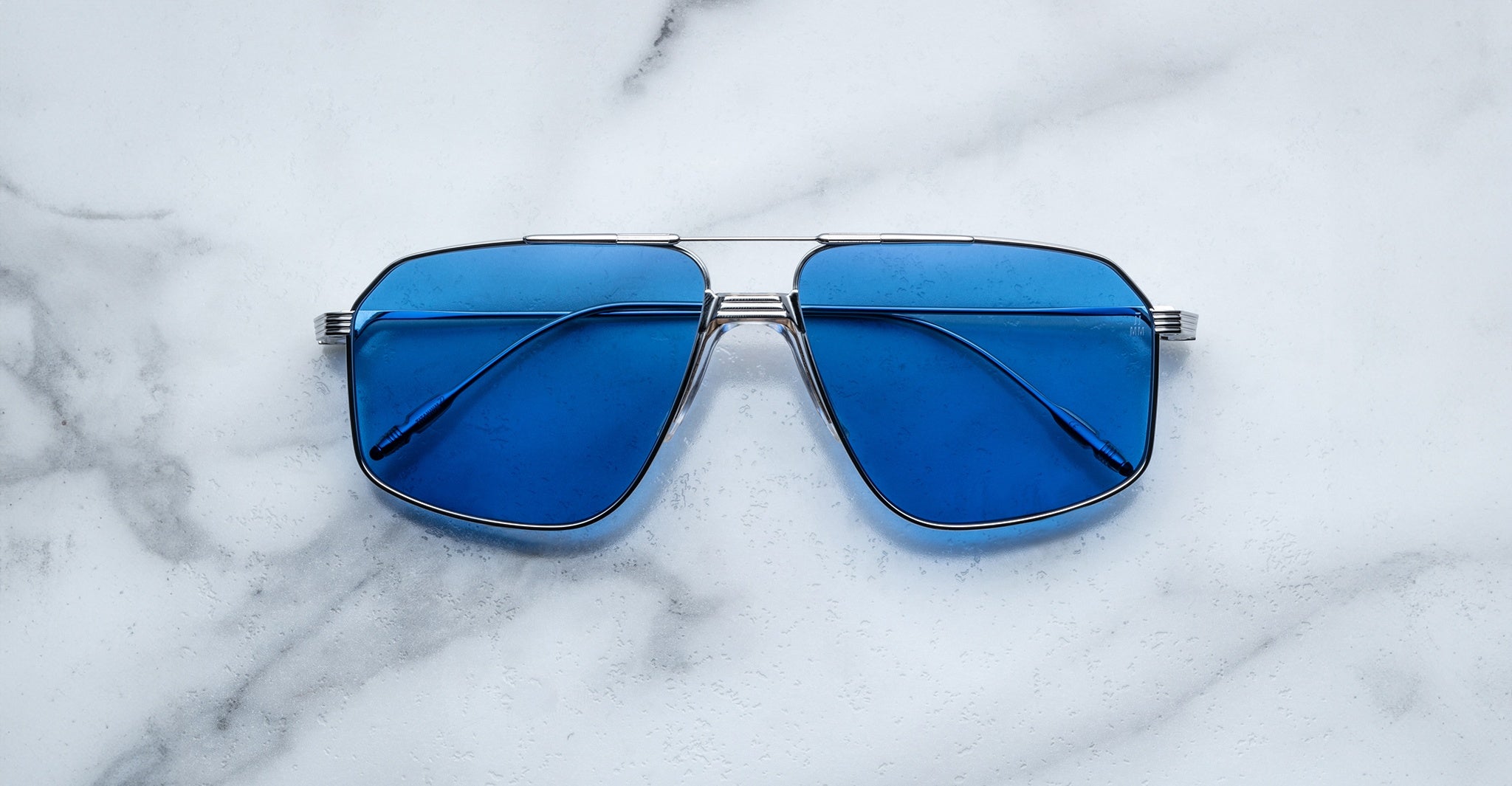 The Jacques Marie Mage Jagger sunglasses by Jacques Marie Mage feature oversized aviator frames in silver beta titanium with blue-tinted lenses, displayed on a white marble surface.