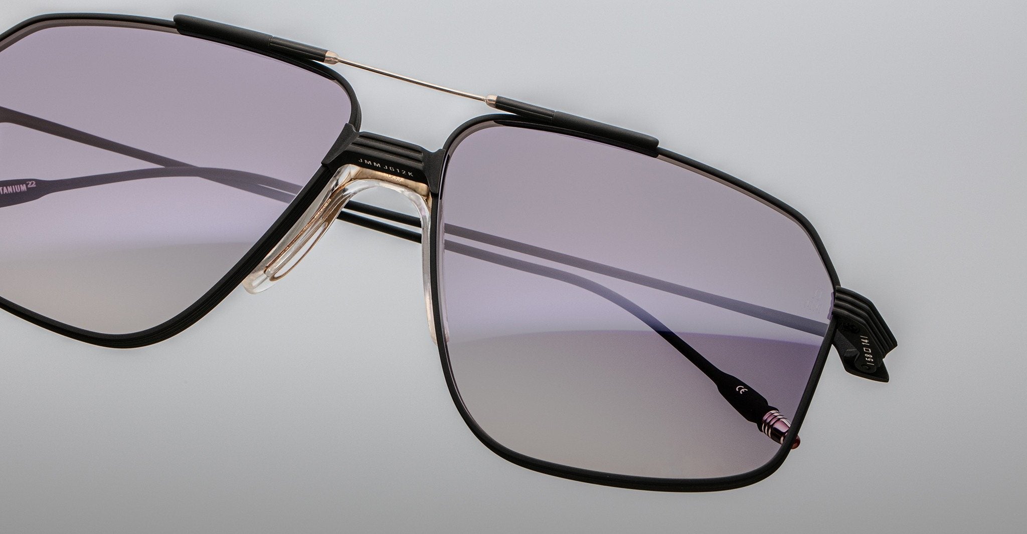 Close-up of Jacques Marie Mage Jagger oversized aviator sunglasses by Jacques Marie Mage, with a thin black and gold beta titanium frame and light purple tinted lenses, set against a plain light background.