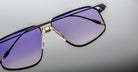 Close-up of Jacques Marie Mage Jagger sunglasses by Jacques Marie Mage, with oversized black frames, beta titanium accents, and gradient purple lenses on a light grey background.