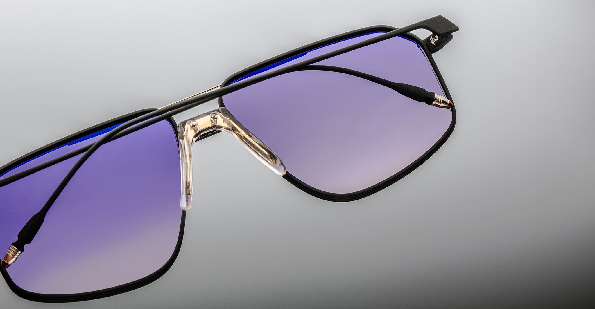 Close-up of Jacques Marie Mage Jagger sunglasses by Jacques Marie Mage, with oversized black frames, beta titanium accents, and gradient purple lenses on a light grey background.