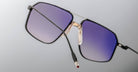 Close-up of Jacques Marie Mage Jagger sunglasses featuring oversized square purple-tinted lenses, black frames, and gold accents in premium beta titanium by Jacques Marie Mage, displayed against a neutral background.