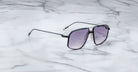 The Jacques Marie Mage Jagger, a pair of oversized aviator sunglasses by Jacques Marie Mage with black frames and gradient lenses, rests on a white marble surface.