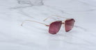 A pair of Jacques Marie Mage Jagger sunglasses, featuring oversized gold frames and large square maroon-tinted lenses, rests on a white marble surface.