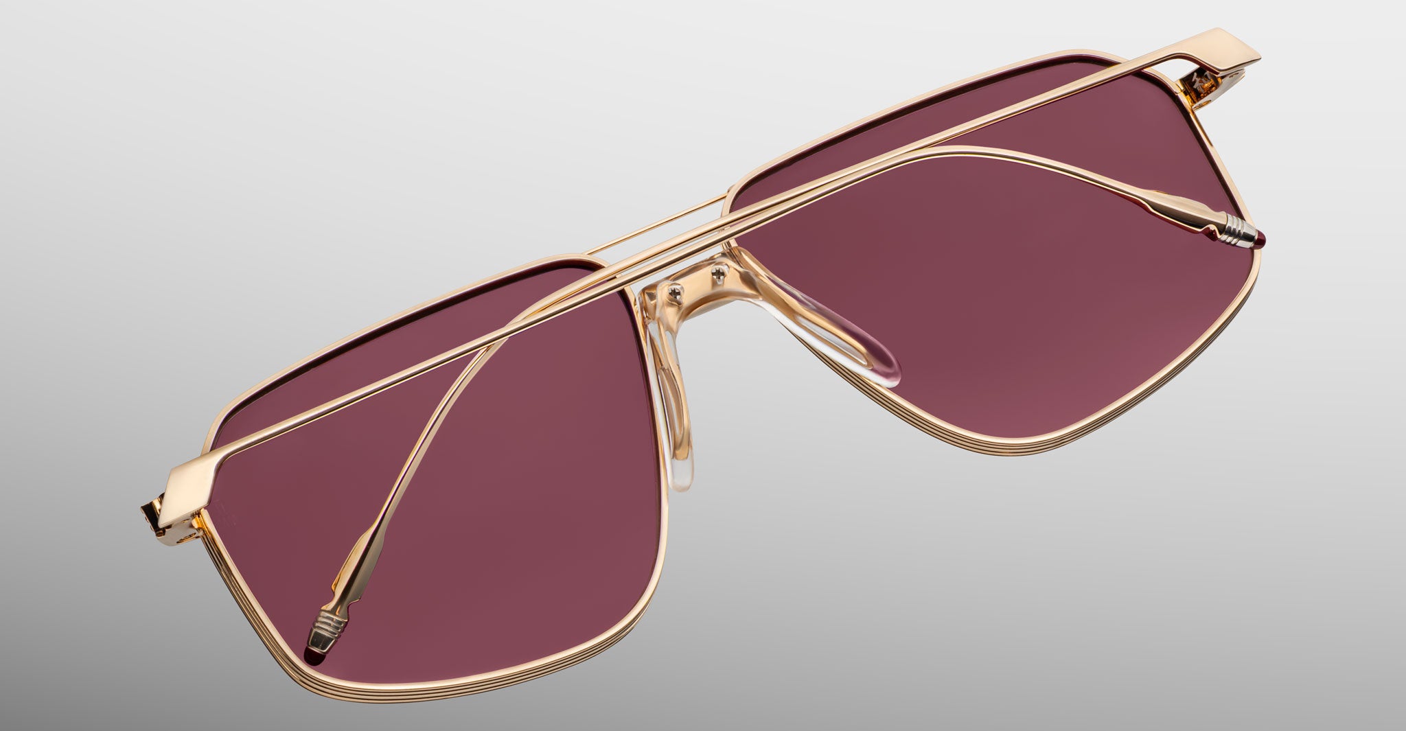 Jacques Marie Mage Jagger sunglasses by Jacques Marie Mage feature a beta titanium frame, gold rims, oversized rectangular rose-tinted lenses, and are shown on a light gray background.