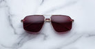 The Jacques Marie Mage Jagger sunglasses by Jacques Marie Mage feature oversized aviator frames in gold with dark red-tinted lenses, displayed elegantly on a white marble surface.