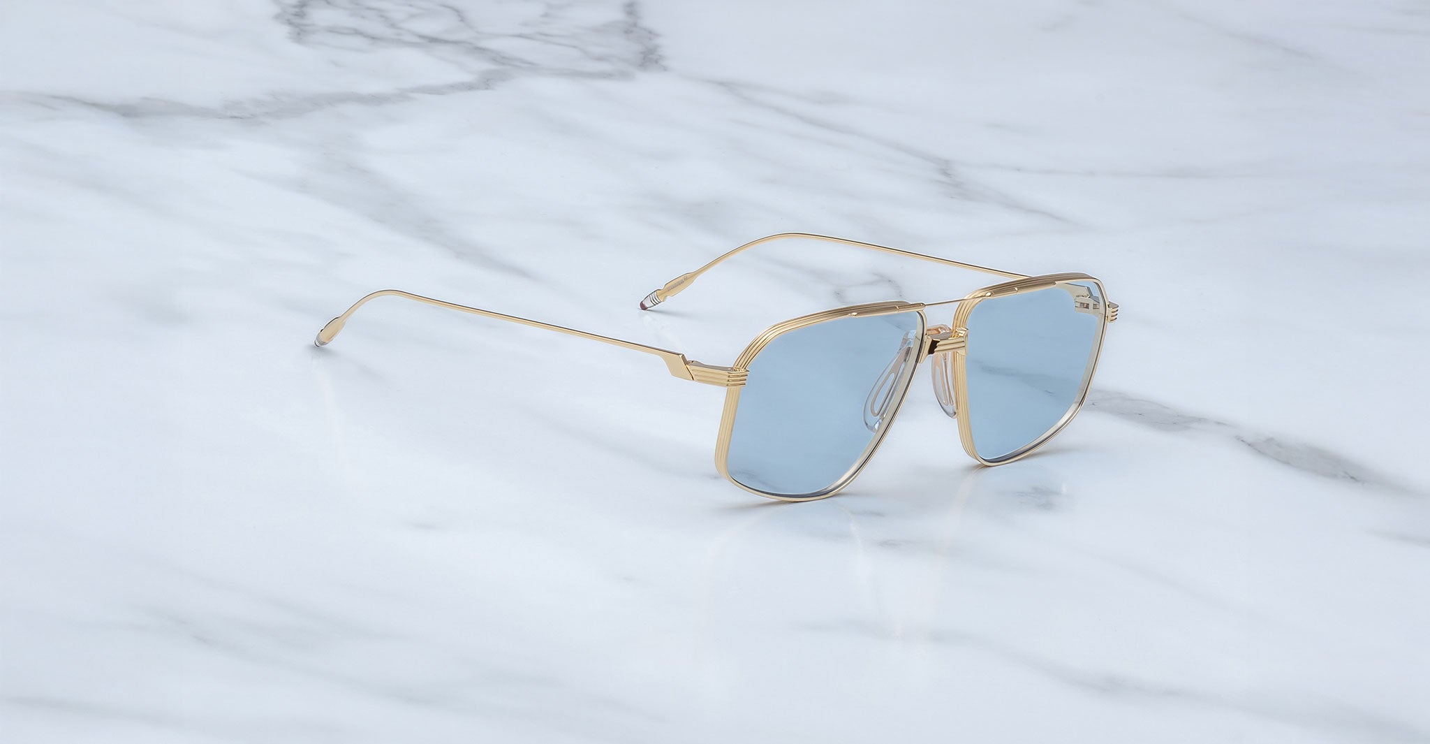 The Jacques Marie Mage Jagger sunglasses by Jacques Marie Mage, featuring gold oversized aviator frames and blue-tinted lenses, are displayed on a white marble surface.
