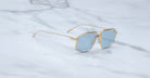 The Jacques Marie Mage Jagger sunglasses by Jacques Marie Mage, featuring gold oversized aviator frames and blue-tinted lenses, are displayed on a white marble surface.