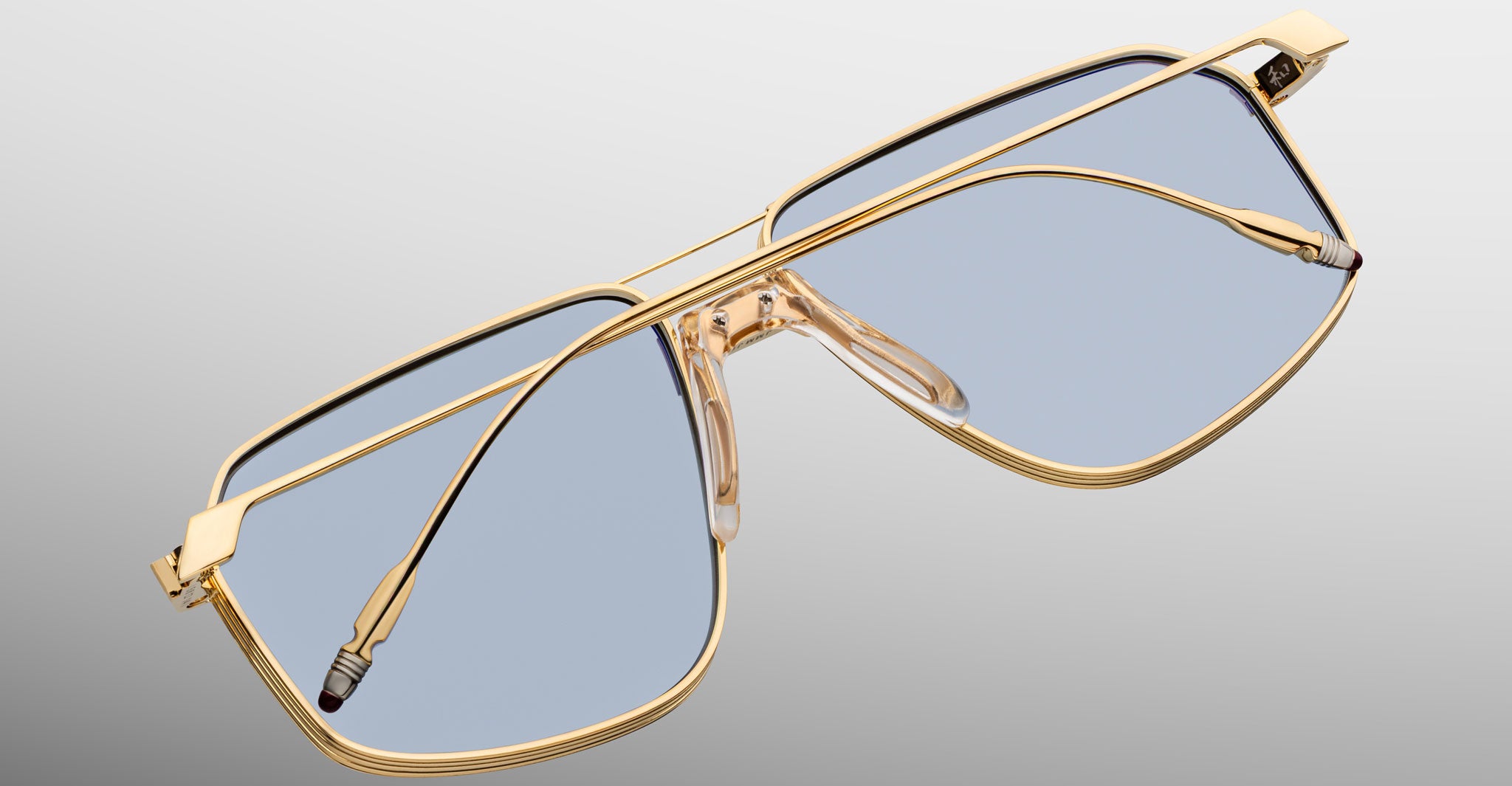 The Jacques Marie Mage Jagger sunglasses feature gold beta titanium frames in an oversized aviator shape with clear blue-tinted lenses, showcased against a gradient gray background to capture their bold essence.