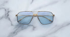 A pair of Jacques Marie Mage Jagger oversized aviator sunglasses with gold frames and blue-tinted lenses by Jacques Marie Mage sits on a white marble surface.