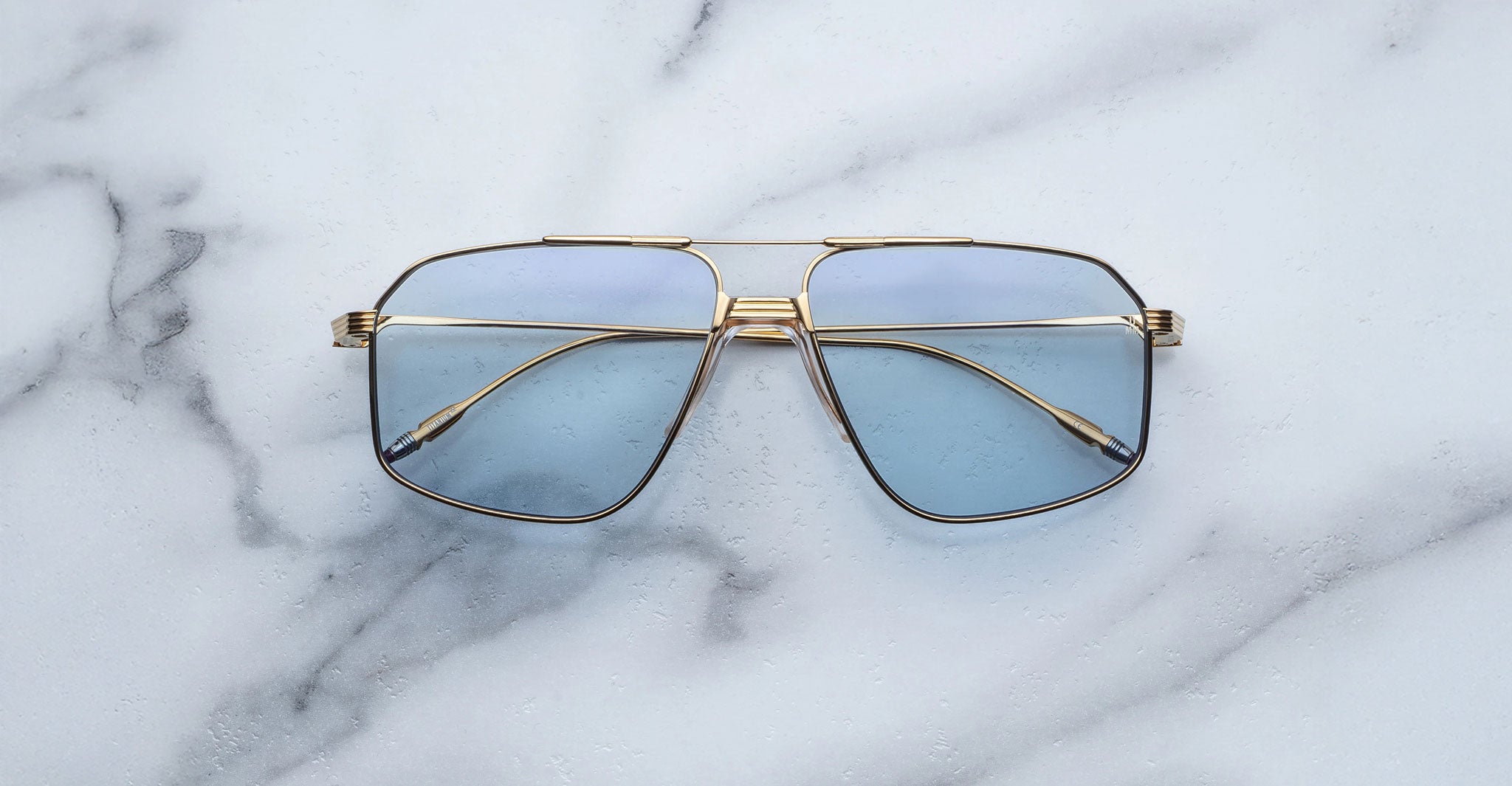 A pair of Jacques Marie Mage Jagger oversized aviator sunglasses with gold frames and blue-tinted lenses by Jacques Marie Mage sits on a white marble surface.