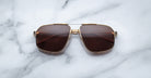 Jacques Marie Mage Jagger sunglasses by Jacques Marie Mage feature gold frames and brown-tinted oversized aviator lenses, displayed on a white marble surface.