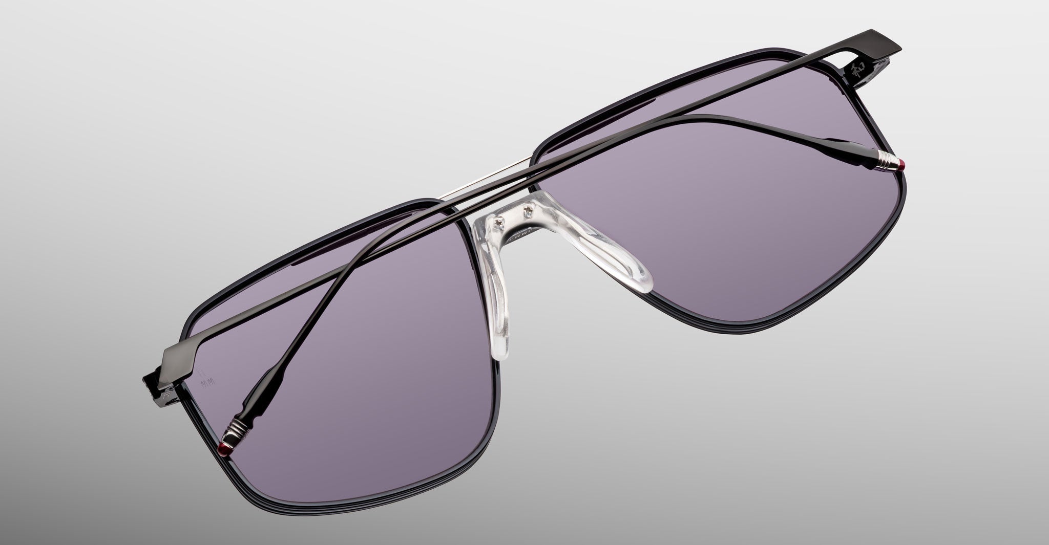 The Jacques Marie Mage Jagger by Jacques Marie Mage is a pair of oversized aviator sunglasses with a black beta titanium metal frame and purple-tinted lenses, showcased on a light gray background.